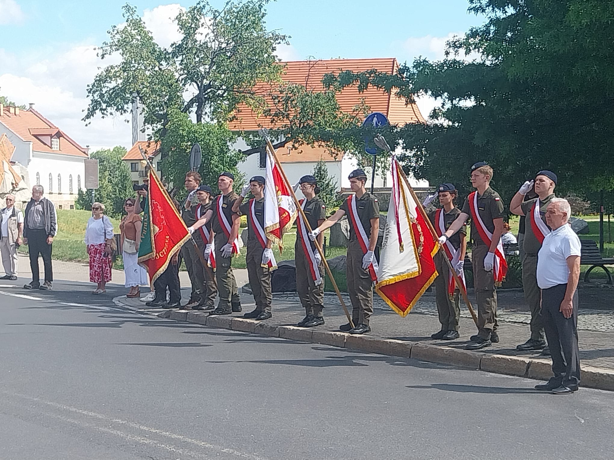 Młodzież w mundurach klas wojskowych trzyma trzy poczty sztandarowe, stojąc na baczność na chodniku podczas uroczystości patriotycznych. Niektórzy kadeci salutują. W tle widoczni są zgromadzeni mieszkańcy, drzewa oraz budynki.
