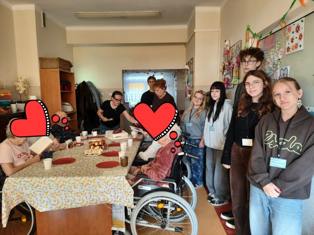 Młodzi wolontariusze i opiekunowie podczas spotkania z seniorami przy stole z poczęstunkiem