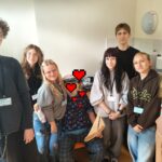 Uśmiechnięta grupa wolontariuszy z identyfikatorami pozuje do zdjęcia z mężczyzną w kapitańskiej czapce i koszulce w żaglówki, który trzyma model żaglówki. Wszyscy są widoczni