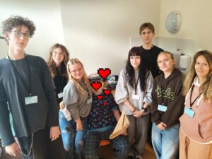 Uśmiechnięta grupa wolontariuszy z identyfikatorami pozuje do zdjęcia z mężczyzną w kapitańskiej czapce i koszulce w żaglówki, który trzyma model żaglówki. Wszyscy są widoczni