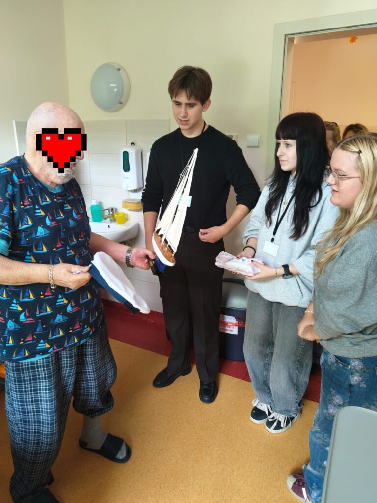 W sali placówki opiekuńczej, starszy mężczyzna (z twarzą zasłoniętą czerwonym sercem pikselowym), ubrany w koszulkę ze wzorem żaglówek, trzyma małą papierową łódkę. Obok niego stoi młody mężczyzna trzymający duży model żaglowca. Dwie młode kobiety patrzą na model. W tle widać zlew