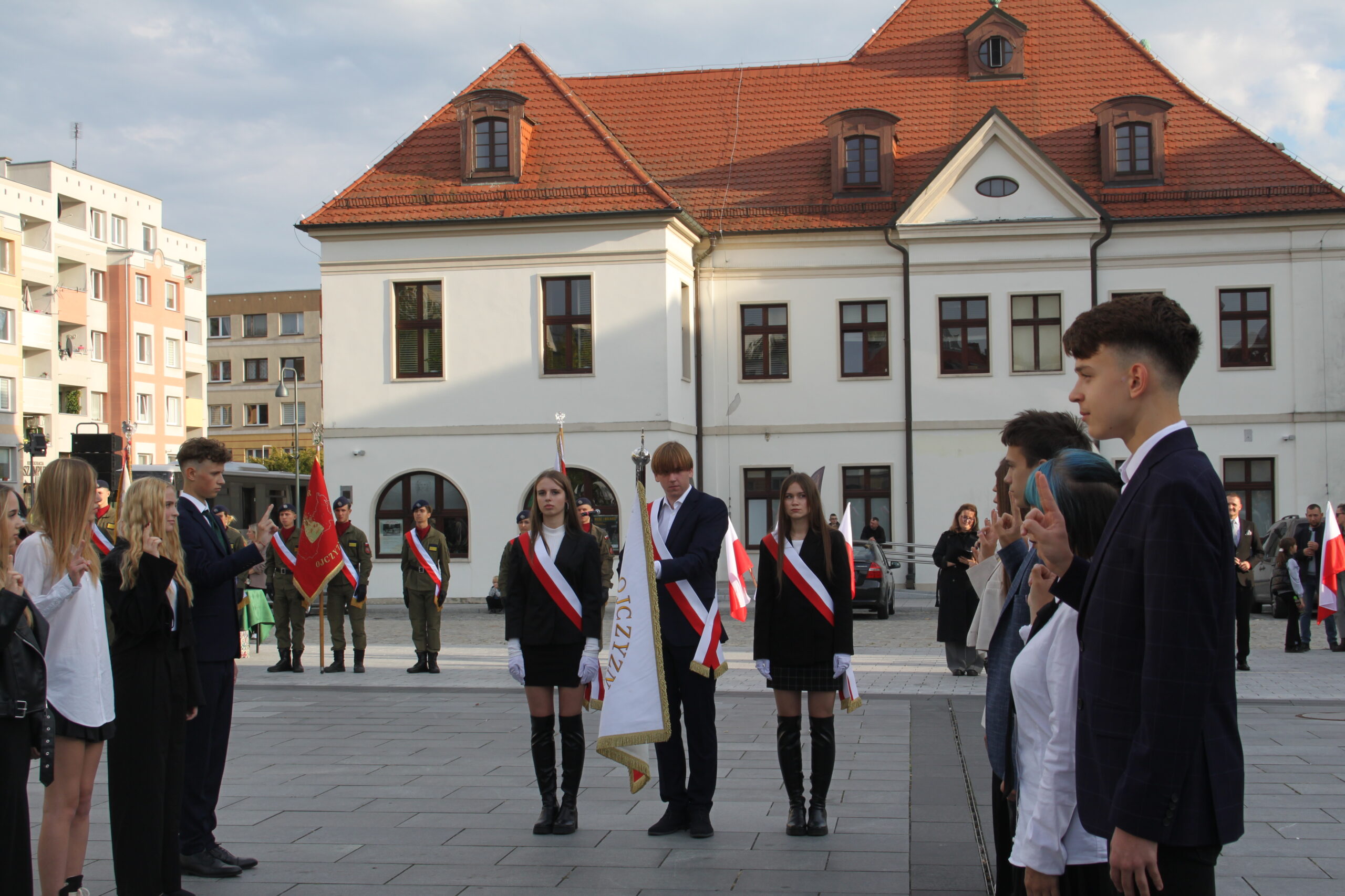 Uczniowie i poczet sztandarowy uczestniczą w oficjalnej uroczystości szkolnej na placu miejskim.