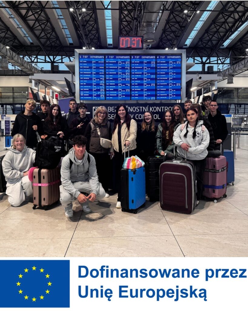 Grupa młodzieży z opiekunami pozująca z bagażami na lotnisku pod tablicą odlotów; na dole grafiki znajduje się napis: Dofinansowane przez Unię Europejską
